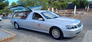 Silver Signature Funeral Services hearse with glass side panels and floral arrangement, parked in cemetery grounds for a dignified funeral service.